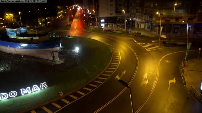 A wet, nighttime roundabout with a model boat in the center is surrounded by buildings and roads with cars driving on them.