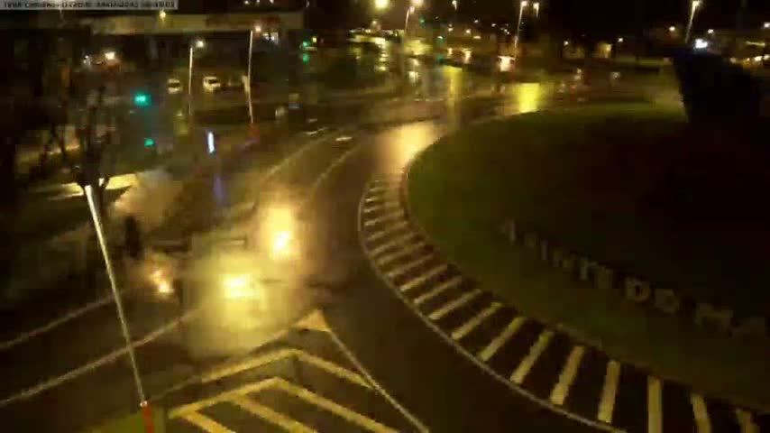 A wet, nighttime scene shows a roundabout with crosswalks and streetlights illuminating a road.