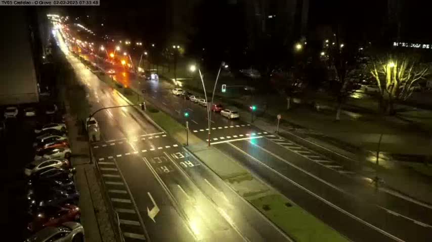 A wet, nighttime street scene shows several cars driving on a multi-lane road that intersects with a crosswalk, and a line of parked cars along the side of the road.