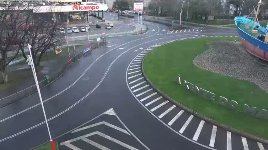 A wet roundabout with a large model boat in the center is surrounded by roads, parking lots, and trees on an overcast day.