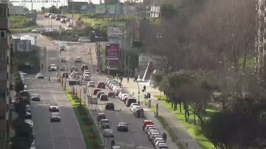 A sunny day shows a multi-lane road with light traffic, lined with trees and buildings.
