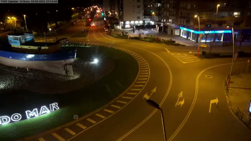 A nighttime aerial view shows a roundabout with a model boat and signage in the center, surrounded by streets and buildings under streetlights.