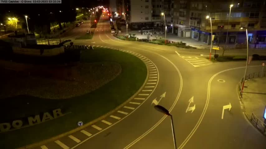 An empty roundabout at night, illuminated by streetlights, is next to a partially visible ship and a row of buildings.