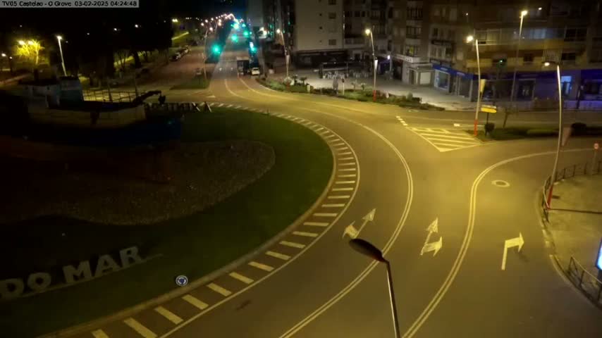 An empty roundabout at night, illuminated by streetlights, is situated near a waterfront with a boat visible in the lower left corner.