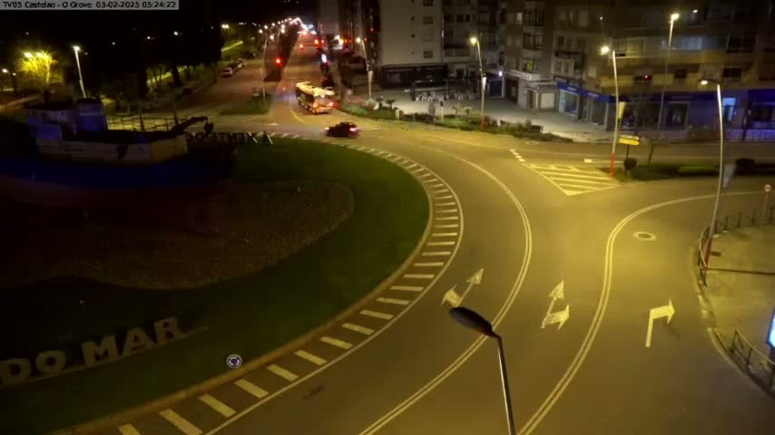 A nighttime view of a roundabout with a partially visible boat-shaped structure beside it, with a few vehicles on the roads and buildings in the background.