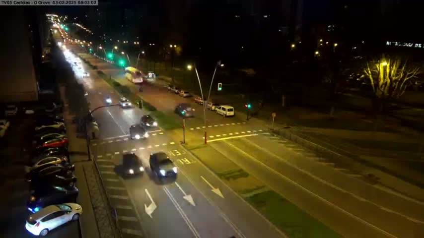 A nighttime, brightly lit street scene shows multiple cars driving in both directions on a multi-lane road, with parked cars along the side and a few pedestrians visible in the distance.