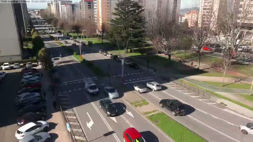 A sunny day reveals a high-angle view of a city street with cars driving on a multi-lane road, surrounded by buildings and trees in a park-like setting.