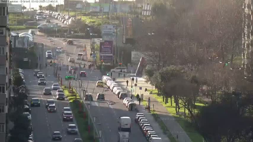 A sunny day reveals a multi-lane road with numerous cars moving in both directions, lined with buildings and trees, and a park-like area with pedestrians.