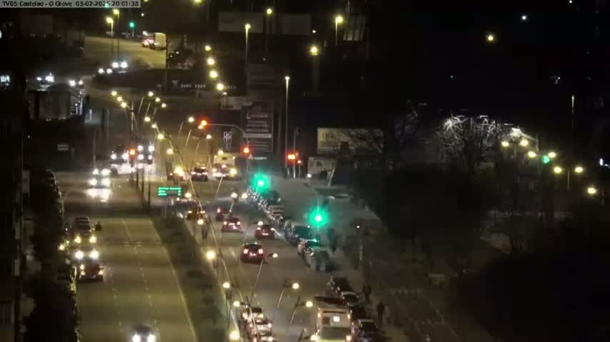 A nighttime view of a multi-lane roadway with cars driving in both directions, under streetlights, and next to a dark, tree-lined area.