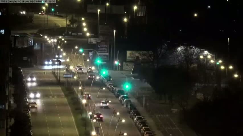A nighttime, high-angle view of a multi-lane road with cars driving in both directions, lined with streetlights and bordered by buildings and trees.
