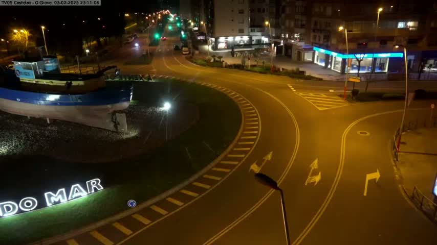 At night, a roundabout features a model fishing boat and signage, surrounded by streets and buildings under artificial light.