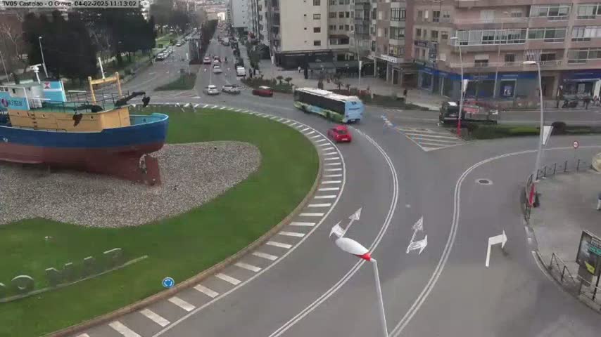A blue and yellow fishing boat sits in a grassy roundabout, surrounded by a curving road with several cars and a bus, under a mostly clear sky.
