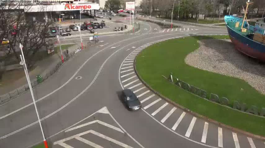 A dark-colored car drives through a roundabout in a city, with a large boat sculpture visible nearby on a sunny day.
