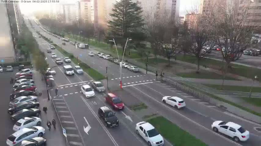 A sunny afternoon view of a multi-lane road with cars driving in both directions, passing through a city intersection with a grassy area and parked cars along the sides.