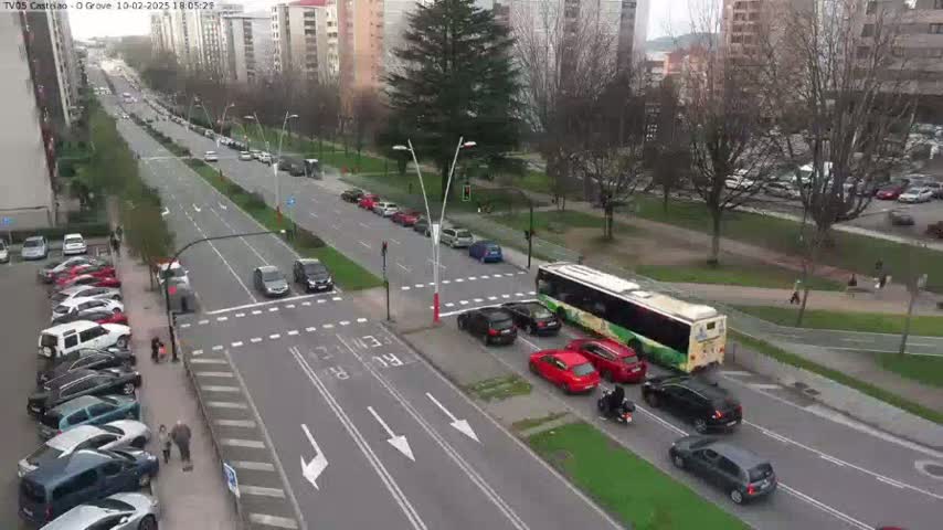 A high-angle view of a city street on a partly cloudy day, showing multiple cars and a bus stopped at an intersection with a parking lot full of cars visible to the left.