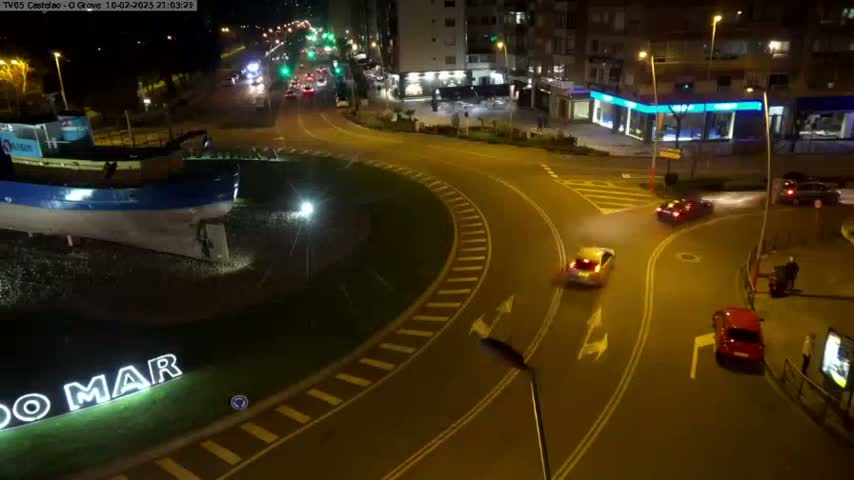 At night, under light rain, several cars navigate a roundabout near a large boat statue in a town with buildings illuminated.
