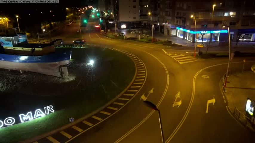 A nighttime, slightly wet roundabout features a large boat sculpture at its center, surrounded by streets and buildings under artificial lighting.