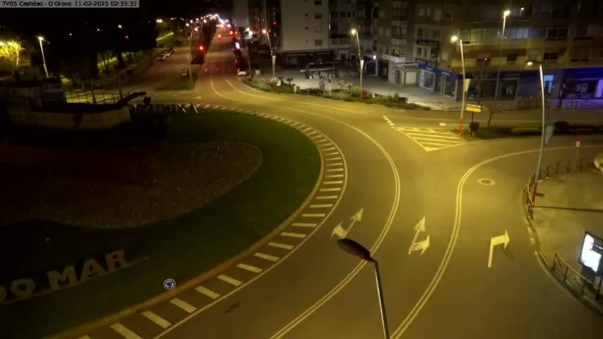 A nighttime aerial view shows a mostly empty roundabout and intersecting roads illuminated by streetlights, with a large dark object resembling a boat visible in the lower left corner.