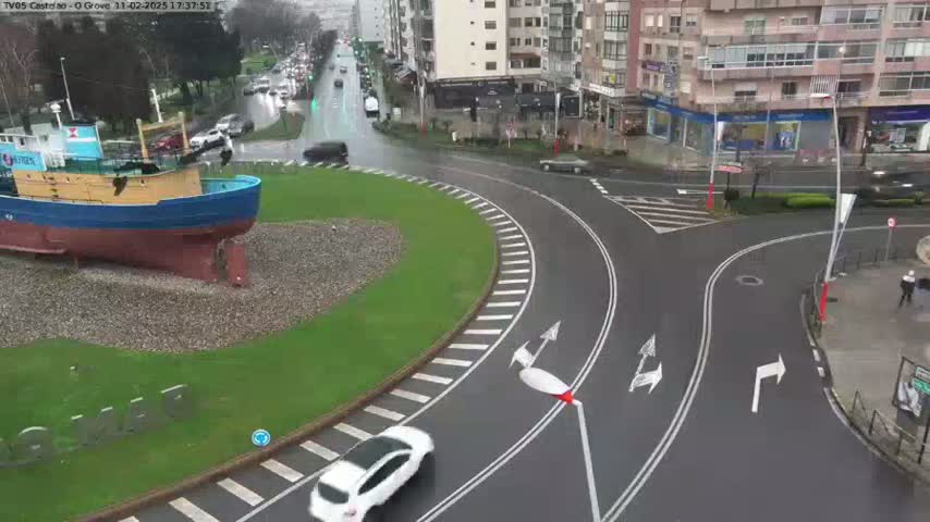 A large blue and brown boat sits on a grassy area next to a wet roundabout with several cars and buildings in the background.