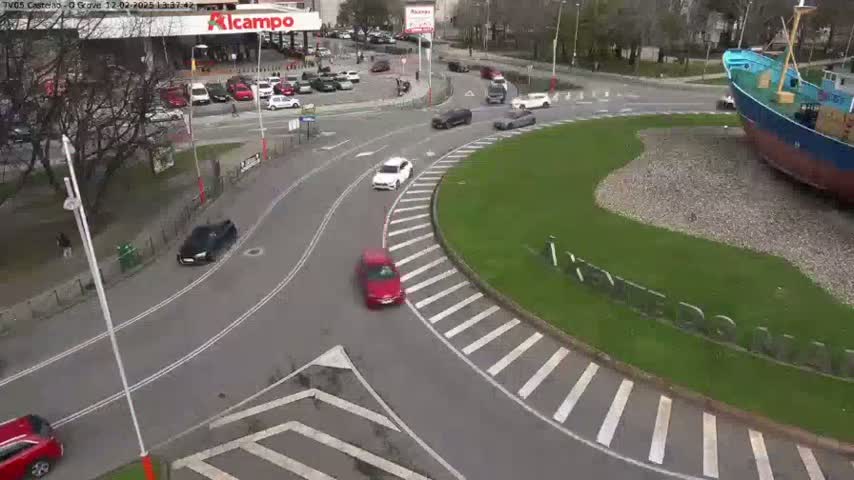Several cars are driving around a roundabout next to a large ship on a partly sunny day.