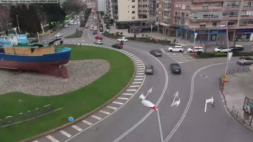 A large, blue-hulled boat sits in a grassy roundabout surrounded by a multi-lane roadway with several cars driving on a partly cloudy day.