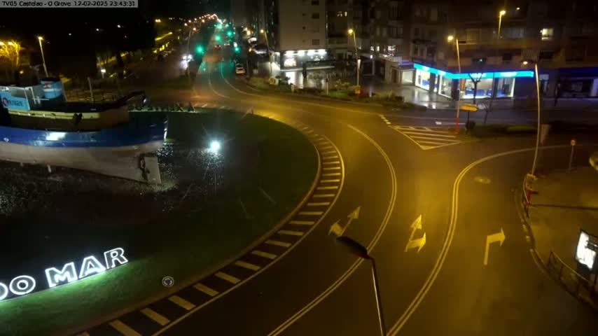 A wet, nighttime roundabout with a boat statue in the center, surrounded by buildings and streets with minimal traffic.