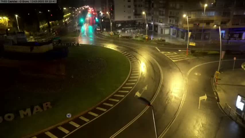 A wet roundabout and intersecting roads at night, illuminated by streetlights, with a few vehicles visible in the distance.