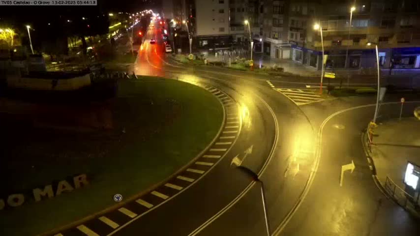 A wet roundabout at night, illuminated by streetlights, with a few vehicles visible in the distance.