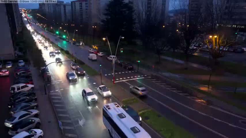 A wet, nighttime street scene shows multiple cars driving on a multi-lane road that passes through an urban area with buildings and trees.