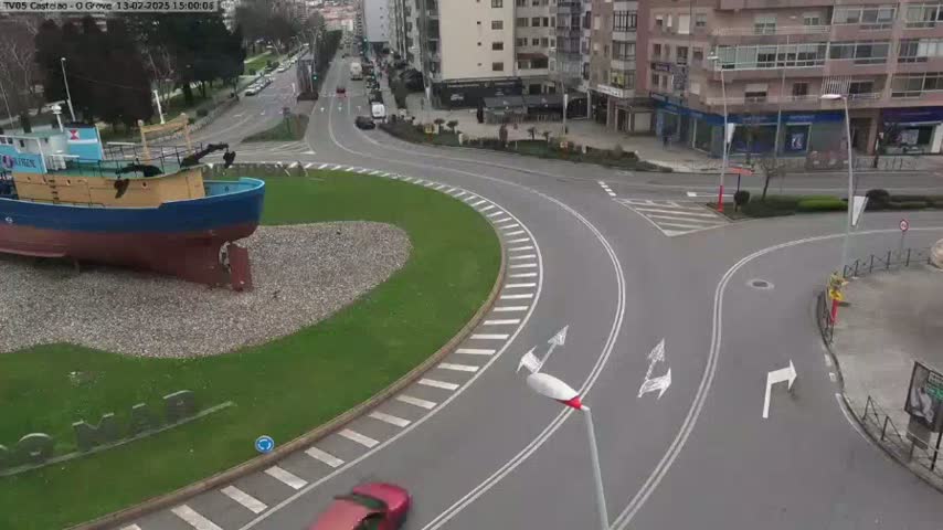 A large, grounded ship sits in a grassy roundabout at the center of a curving road intersection, under an overcast sky.