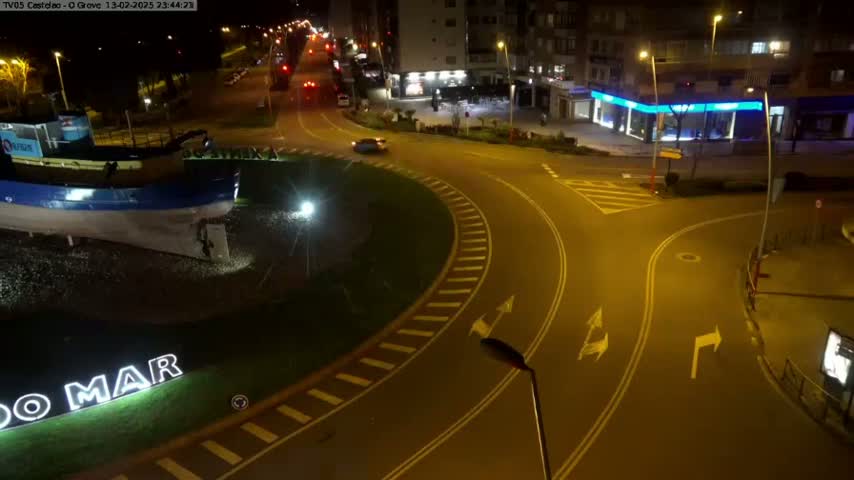A nighttime aerial view shows a roundabout with a large boat sculpture at its center, near buildings and roads with cars driving on them.