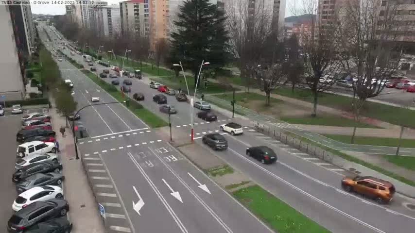 A partly sunny day reveals a multi-lane road with moderate traffic, lined with parked cars and bordered by buildings and trees.