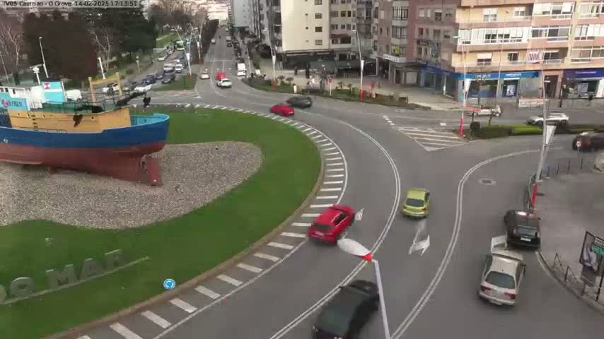 A large model of a blue and yellow fishing boat sits in a grassy roundabout at the center of a multi-lane roadway with several cars navigating the curved lanes on a partly sunny day.