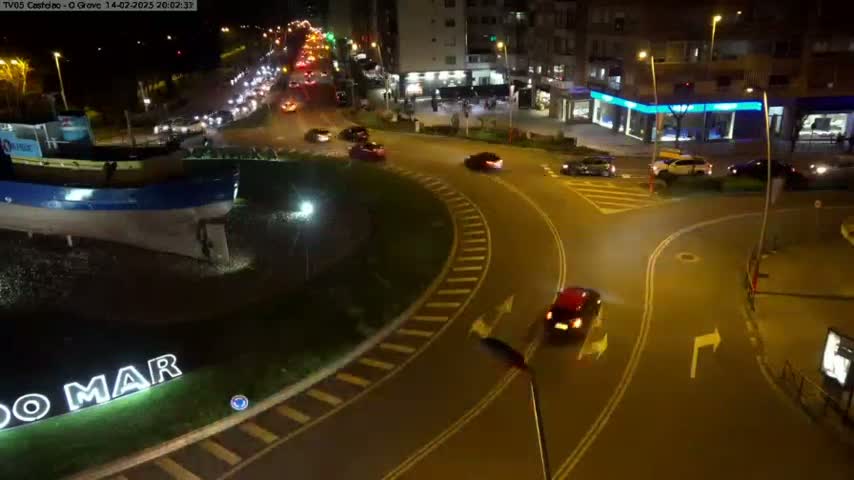 At night, a roundabout with several cars driving around it, near a large boat statue and buildings.