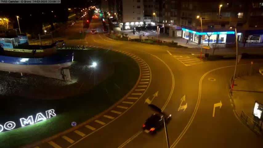 At night, a dark-colored vehicle drives through a roundabout near a large model of a boat and illuminated signage, next to buildings under streetlights.