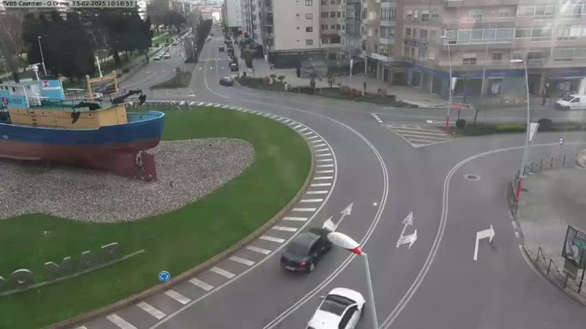 A large fishing boat sits in a grassy roundabout next to a curving road with several cars, in what appears to be an overcast day.