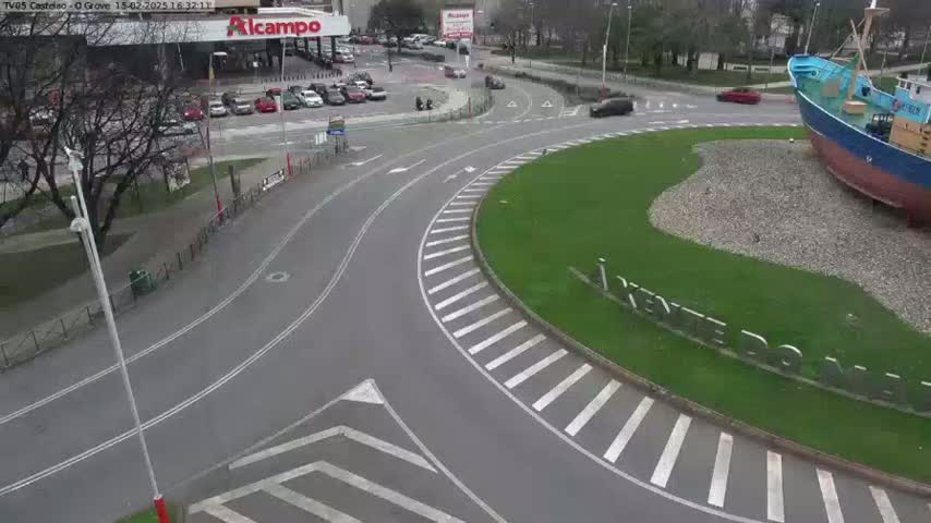 A roundabout with a large boat sculpture in the center, surrounded by a grassy area, with several cars parked nearby and a few driving on an overcast day.