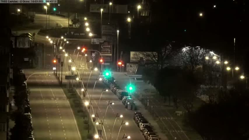 A nighttime view of a multi-lane road with parked cars on either side, illuminated by streetlights, and some traffic moving along the road.