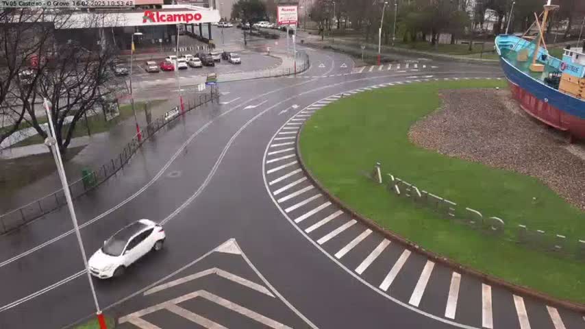 A wet, curving roadway with crosswalks encircles a grassy area where a large ship sits, with a parking lot and buildings visible in the background.