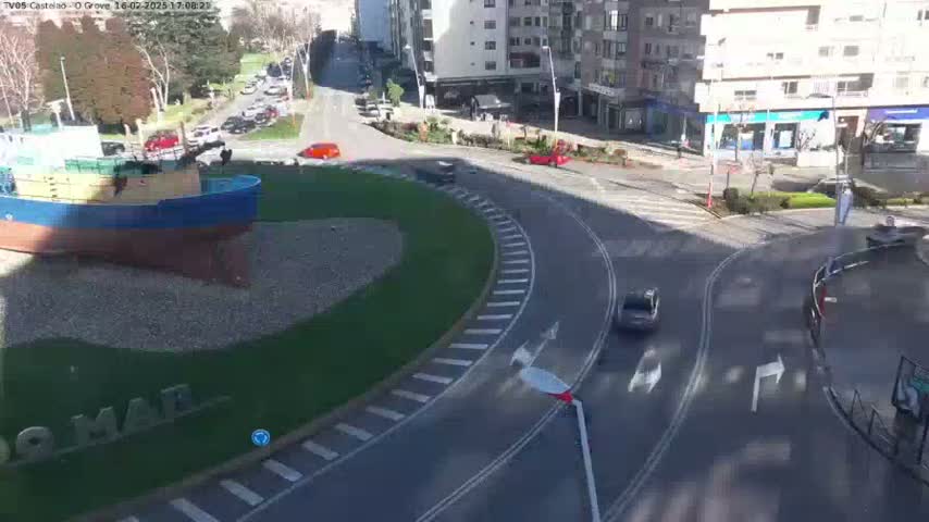 A model of a fishing boat sits in a grassy area next to a roundabout with several cars driving on a sunny day.