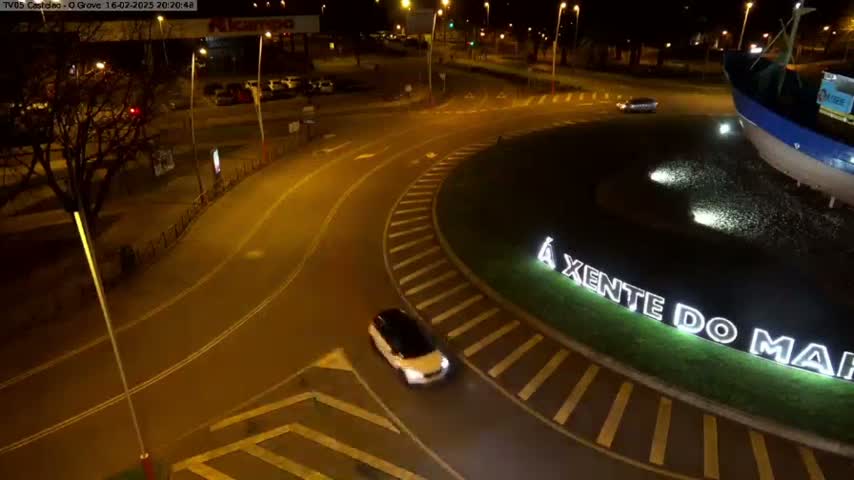 A white car drives around a roundabout at night, illuminated by streetlights, near a large model of a fishing boat.