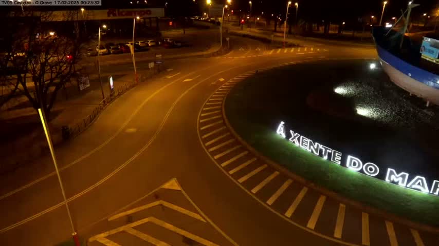 An aerial nighttime view shows a roundabout with a large boat sculpture and illuminated signage in the center, surrounded by roadways and parking lots under artificial light.
