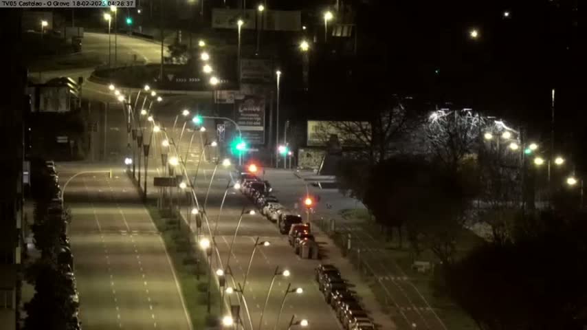 A nighttime view of a multi-lane road with parked cars and streetlights, bordered by trees and buildings.
