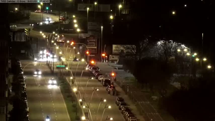 A nighttime, high-angle view of a multi-lane road with cars driving in both directions, lined with streetlights and bordered by dark trees and buildings.