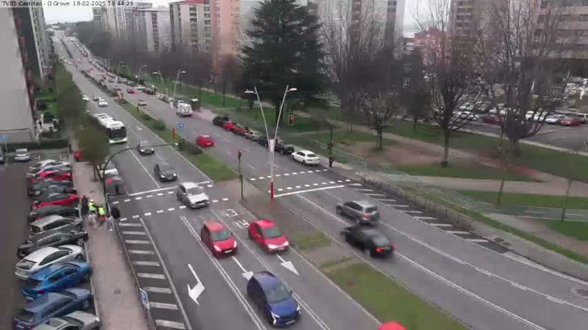 A city street scene on an overcast day shows several cars driving in both directions on a multi-lane road, with parked cars lining the street, and a pedestrian crossing.
