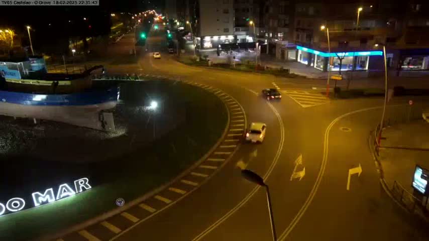 At night, a roundabout with a few cars is illuminated, featuring a large boat sculpture in the center.