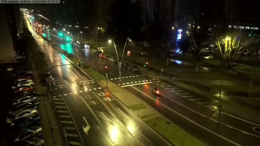 A rain-slicked city street at night shows cars driving in both directions, with parked cars lining the side of the road, and a lone motorcyclist in the middle of the road.
