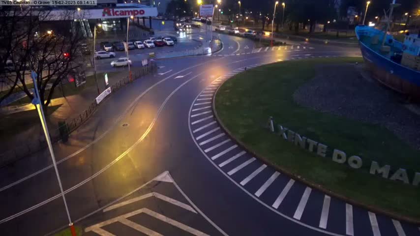A wet roundabout at night is surrounded by parked cars, streetlights, and a large model of a fishing boat.