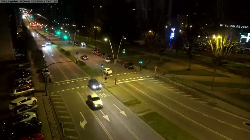 A nighttime aerial view of a multi-lane road with cars driving in both directions, intersecting with a crossroad, surrounded by parked cars and streetlights illuminating a park-like area.