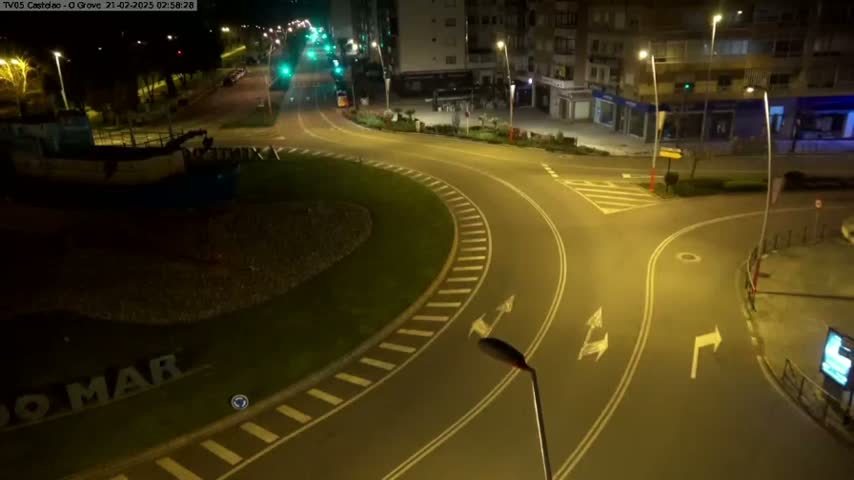 A mostly empty roundabout at night, illuminated by streetlights, with a boat visible in the background.